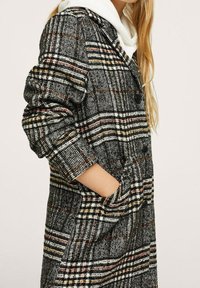 Cappotto a scacchi in nero, bianco e colori, con colletto a revers, bottoni frontali e tasche laterali. Il materiale ha un aspetto texturizzato e tessuto.