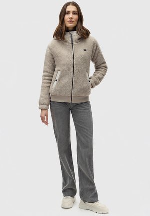 Veste en polaire beige avec un col haut, fermeture éclair, poches latérales et poignets côtelés. Associée à un pantalon large gris et des baskets.