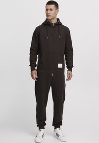 Brown zip-up jumpsuit made of soft fabric, featuring a hood, front pockets, and a label on the left side. Model wears white sneakers.