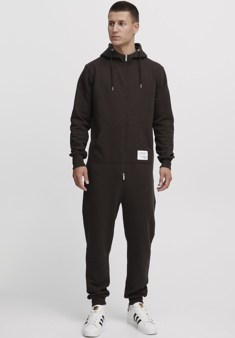 Brown zip-up jumpsuit made of soft fabric, featuring a hood, front pockets, and a label on the left side. Model wears white sneakers.