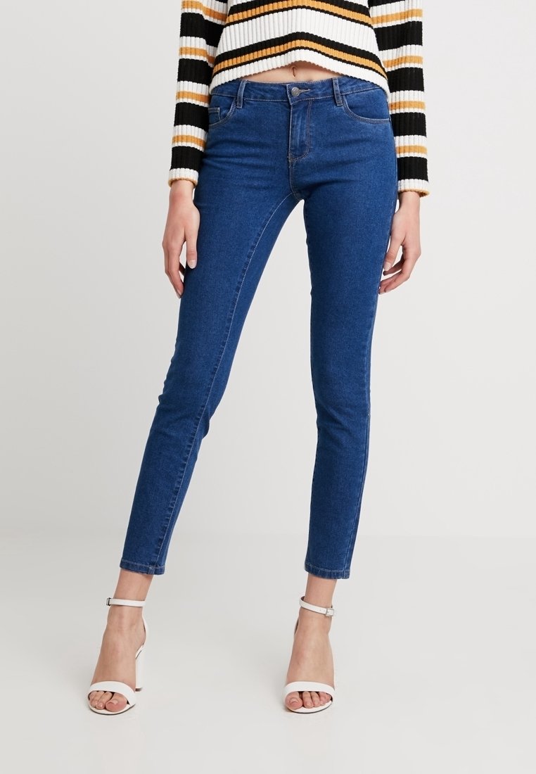 Jeans skinny de mezclilla azul con una cintura de altura media, que presentan una textura suave y un diseño clásico de cinco bolsillos, combinados con sandalias de tacón alto blancas.