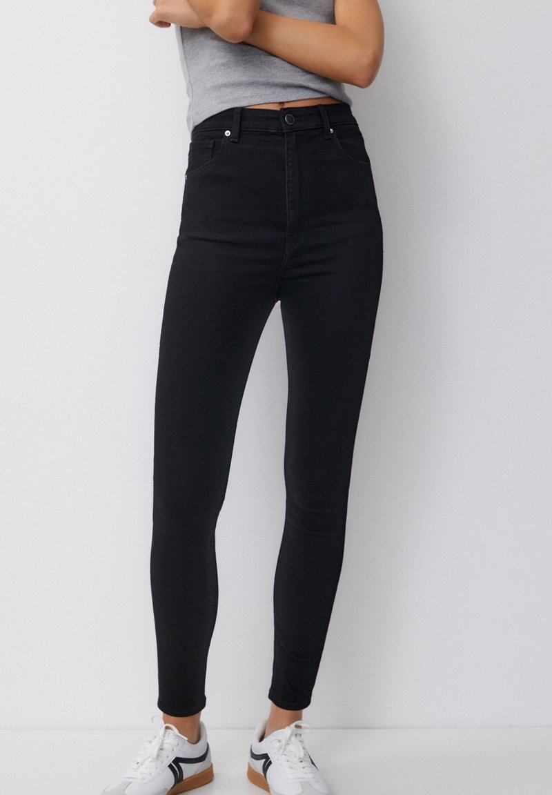 Jeans skinny de tiro alto en denim negro con una textura suave, que cuentan con bolsillos frontales y un cierre de botón, combinados con zapatillas blancas.