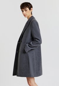 Cappotto in misto lana grigio con una vestibilità rilassata, caratterizzato da un colletto a scowl, due tasche frontali e lunghezza a metà coscia. Tessuto morbido.
