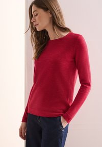Rotes Langarmshirt mit strukturiertem geripptem Design und rundem Ausschnitt, kombiniert mit dunkelblauen Jeans. Der Stoff wirkt weich und bequem.