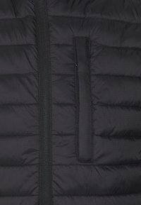 Svart vadderad jacka med en vertikal dragkedja och en liten dragkedjeficka. Tyget har en mjuk textur med ett subtilt rutmönster.