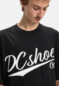 Černé bavlněné tričko s velkým bílým nápisem "DC shoe co" a zakřiveným podtržením, s klasickým výstřihem do kruhu, krátkými rukávy a uvolněným střihem.