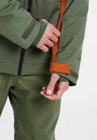 Wasserdichte grüne Jacke mit orangefarbenen Akzenten, ausgestattet mit einer Reißverschlusstasche und verstellbaren Bündchen mit Klettverschlüssen. Weiches, strukturiertes Material.