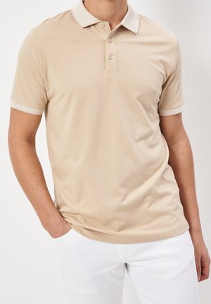 Mann trägt beige Polohemd mit kurzen Ärmeln und weißem Rand sowie weiße Hose, steht mit einer Hand in der Tasche vor einfachem Hintergrund.