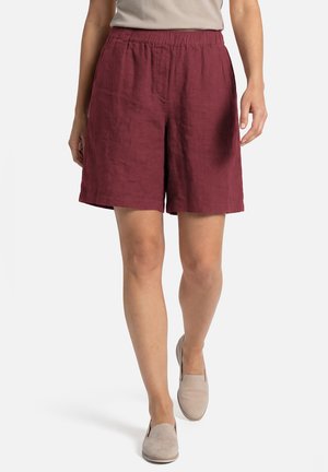 Personne portant un short ample bordeaux et des chaussures beiges à enfiler, marchant devant un fond blanc uni.