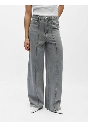 Femme portant un jean gris taille haute à jambes larges avec des détails de couture avant et des chaussures blanches à bout pointu, debout devant un fond uni.