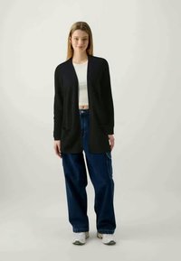 Black cardigan with open front, long sleeves, and side pockets, worn over a white top, paired with loose dark blue jeans and sneakers.