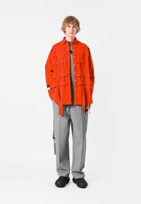 Chaqueta roja brillante de gran tamaño con bolsillos de parche, camiseta gris, pantalones gris claro y zapatos negros texturizados con suela gruesa.