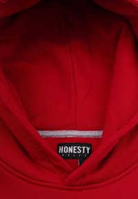 Punainen huppari pehmeällä pinnalla, jossa on harmaa vuorattu huppu. Etiketissä lukee "HONESTY RULES" mustalla taustalla.
