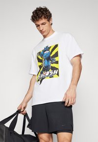 White cotton t-shirt featuring a blue superhero graphic with a yellow and black striped background; worn with black shorts and holding a bag.
