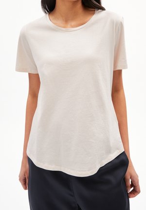 Kobieta w prostym jasnobeżowym t-shircie z krótkim rękawem i okrągłym dekoltem oraz ciemnoniebieskich spodniach, stojąca na białym tle.