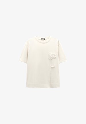 Biała krótki rękaw bawełniana koszulka z teksturowanym wzorem kwiatowym i węzłem po lewej stronie. Luźny krój, prosty okrągły dekolt.