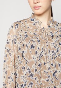 Lätt blus med mönster i beige med blå detaljer, som har krage med ruffles och knytning vid halsringningen. Texturerad ytdesign.