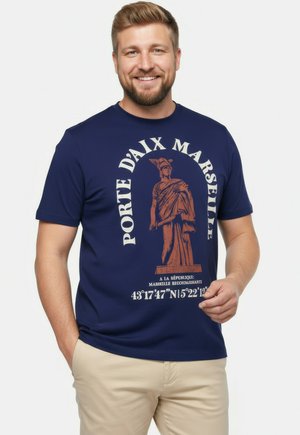 Lächelnder Mann mit kurzem blondem Haar und Bart, der ein marineblaues T-Shirt mit Statue-Grafik und dem Text „Porte d'Aix Marseille“ sowie beige Hose trägt.