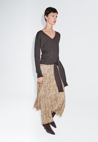 Frau trägt einen dunkelbraunen V-Ausschnitt-Pullover, der an der Taille gebunden ist, einen beige gemusterten Rock und dunkelbraune Stiefel mit Absatz, steht vor einem weißen Hintergrund.