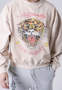 Sudadera beige con un gran gráfico de un tigre rugiente con rayas negras y amarillas. El texto dice "Ed Hardy" y "Los Ángeles".