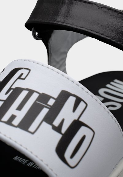 Κοντινό πλάνο λουριού σανδάλιου σε μαύρο και άσπρο χρώμα με έντονο μαύρο κείμενο "Moschino" τυπωμένο σε λευκό δέρμα.