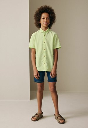 Barn med krøllet hår iført en lysegrøn kortærmet skjorte med knapper, mørke cowboyshorts og brune sandaler, stående indendørs.