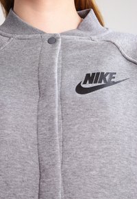 Felpa Nike grigia con chiusura a bottoni, tessuto testurizzato e logo nero sul lato sinistro del petto. Presenta un colletto alto e dettagli a coste.