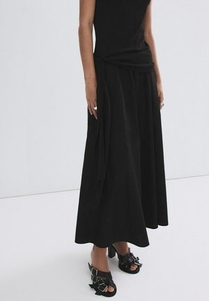 Mujer con vestido negro sin mangas y sandalias negras de tacón con doble hebilla, posando frente a un fondo blanco liso.