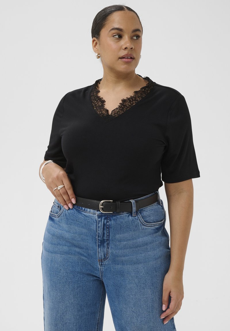 Femme portant un haut noir à col en V orné de dentelle, un jean bleu taille haute avec ceinture noire, un bracelet en argent et des bagues, regardant légèrement sur le côté.