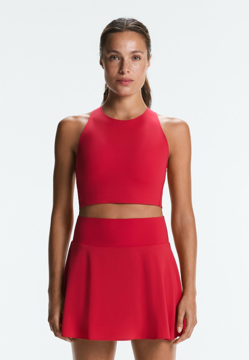 Femme aux cheveux châtain clair attachés, portant un crop top rouge sans manches et une jupe rouge assortie sur fond uni.
