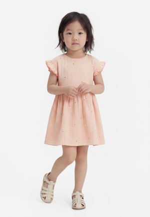 Young girl standing, wearing a light peach knee-length dress with flutter sleeves and beige closed-toe sandals.