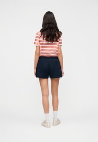Κοντομάνικο ριγέ πουκάμισο σε κόκκινο και άσπρο, συνδυασμένο με σορτς σε ναυτικό μπλε. Το σορτς διαθέτει ελαστική μέση και μία πίσω τσέπη.