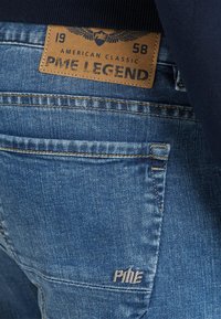 Jeans in denim blu con etichetta in cuoio beige che riporta la scritta "PME Legend" e un design di aquila. Le caratteristiche includono cuciture sulle tasche posteriori e ricamo del logo.
