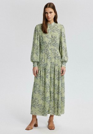Femeie îmbrăcată într-o rochie florală lungă, verde deschis, cu mâneci bufante, cu nasturi în față și sandale cu toc bloc deschis la vârf, stând în fața unui fundal simplu.