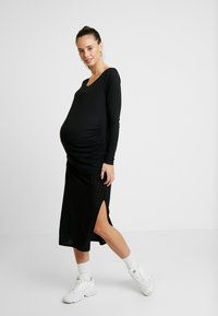 T-shirt de maternidade preta de manga longa com textura canelada; combinada com uma saia preta até ao joelho, com uma fenda lateral, usada com ténis brancos.