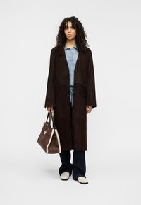 Manteau long en daim marron avec un col cranté et des poches avant, porté sur une chemise rayée bleue et un jean foncé. Porte un sac marron avec des accents en peau lainée.