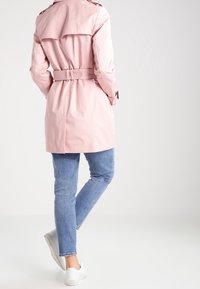 Ljusrosa trenchcoat med knytmidja, knälång design, stor ryggklaff och knappade ärmar, kombinerad med blå jeans och vita sneakers.