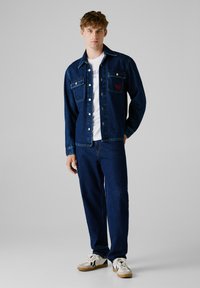 Chaqueta de mezclilla azul índigo con frente abotonado, dos bolsillos en el pecho y camiseta blanca. Combinada con pantalones de mezclilla a juego y zapatillas.