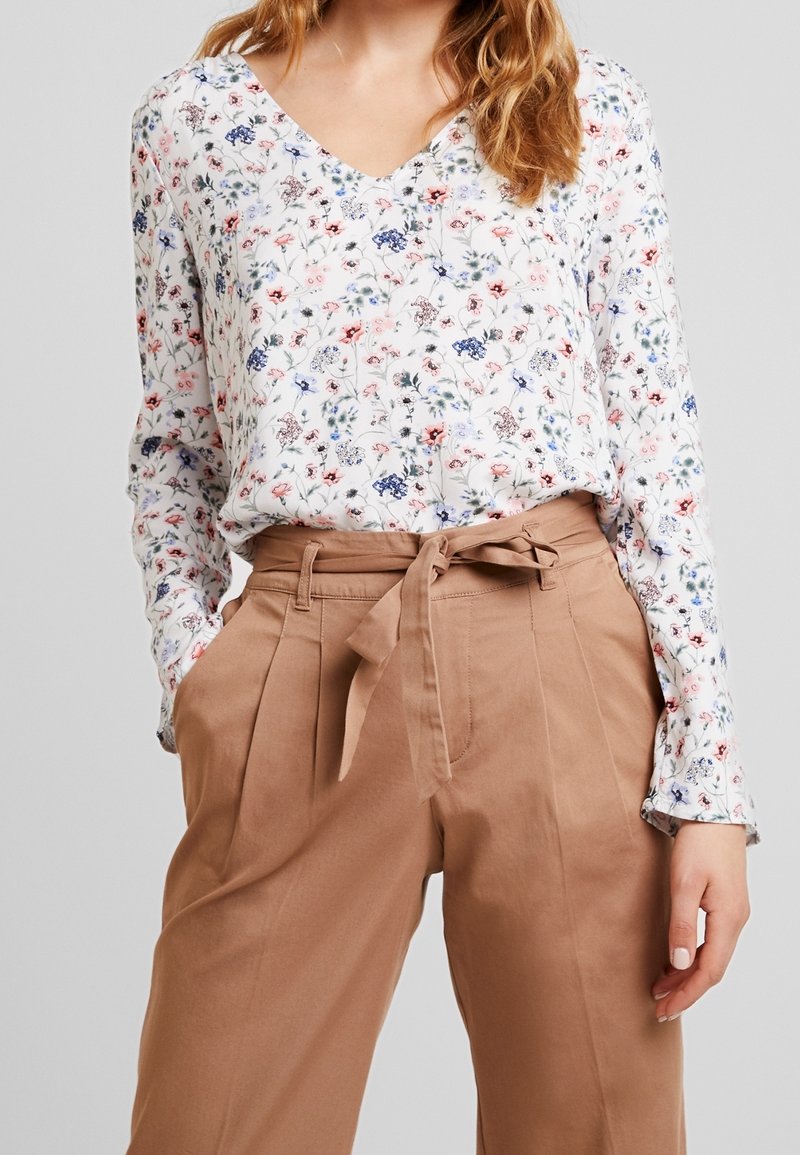 Blusa floral con escote en V, que presenta pequeñas flores multicolores sobre un fondo blanco. Combinada con pantalones de tiro alto de color marrón claro con cinturón de lazo.