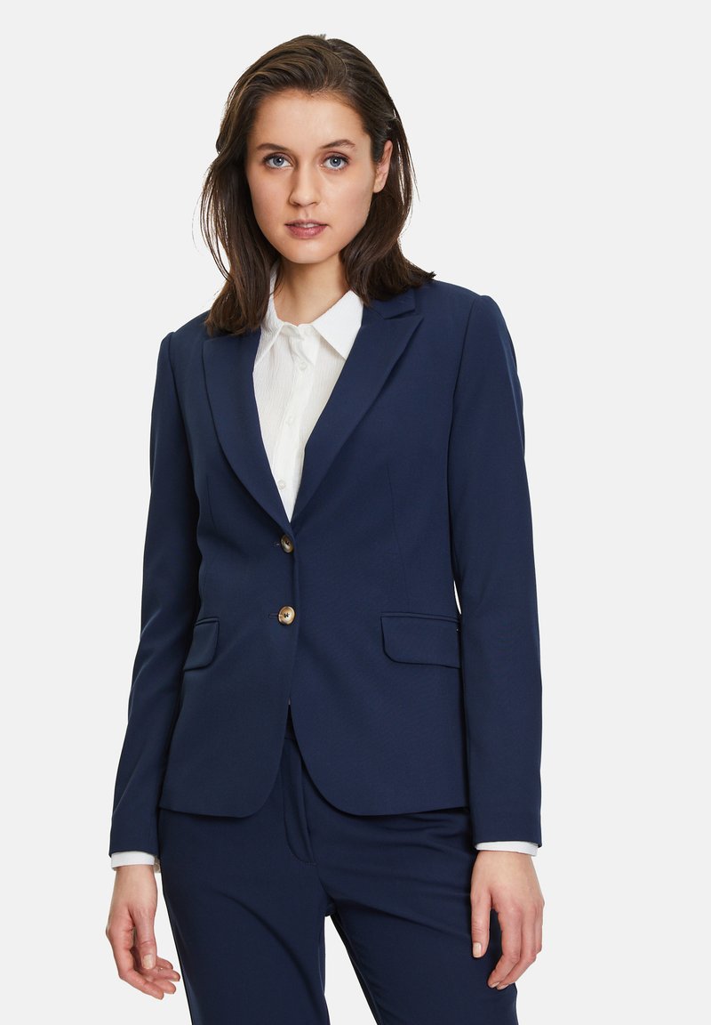 Námořnický modrý blazer se strukturovaným designem, dvěma předními kapsami, zapínáním na jeden knoflík a zlatými prvky. Spárováno s bílou košilí.