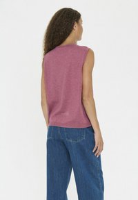 Eine Frau mit lockigem Haar steht mit dem Rücken zur Kamera und trägt ein ärmelloses Oberteil in staubigem Rosé und hochtaillierte Jeans in Blau vor einem schlichten Hintergrund.