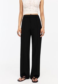 Pantalones de pierna ancha negra con una textura suave, que cuentan con pliegues frontales y un corte ajustado. Combinados con un top corto de encaje de color claro y sandalias negras.