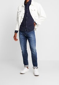 Bílá bunda, tmavě modrá košile na knoflíky, modré džíny a bílé tenisky. Bunda má dvě přední kapsy a knoflíky v odstínu zlata.