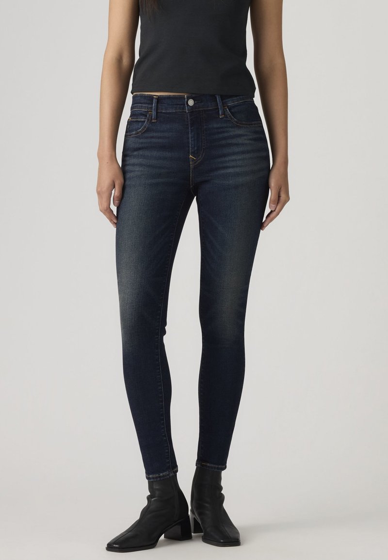 Femeie purtând blugi skinny albastru închis, top negru fără mâneci și cizme negre cu toc la gleznă, stând în fața unui fundal gri.