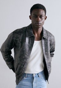 Textureret sort jakke med et reptilhudsmønster, med krave og trykknaplukning, båret over en hvid skjorte og blå jeans.