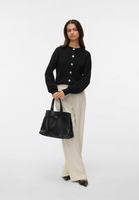 Cárdigan negro con cuello redondo y botones plateados, combinado con pantalones anchos beige y un bolso tote de cuero negro. Texturas suaves.