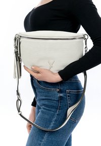 Naine, kes kannab musta pika varrukaga topi ja siniseid teksapükse, hoiab suurt valget perforatsioonidega käekotti, millel on lukk ja tutid.