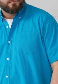 Korte mouw, knoopsluiting overhemd van helder turquoise stof. Heeft een enkele borstzak, lichte textuur en witte knopen.