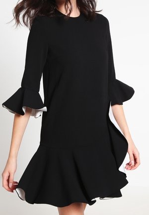 Frau trägt ein schwarzes Kleid mit ausgestellten dreiviertel Ärmeln und einem gerüschten Saum, das weiße Futterdetails aufweist.