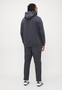 Man with dreadlocks wearing dark gray hoodie and cargo pants, white sneakers with black stripes, standing facing away on plain background.
